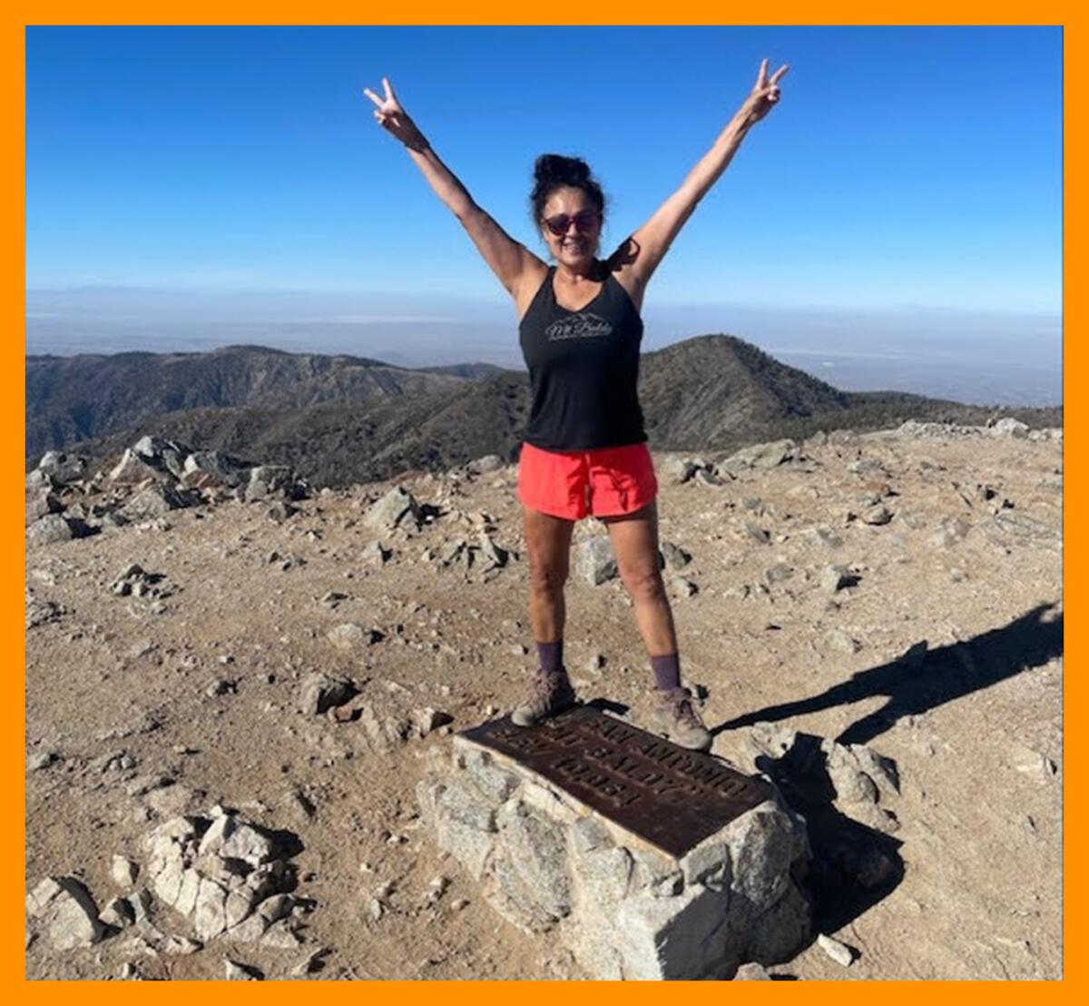 A woman in shorts and a tank top stands with her arms upraised on a mountain.