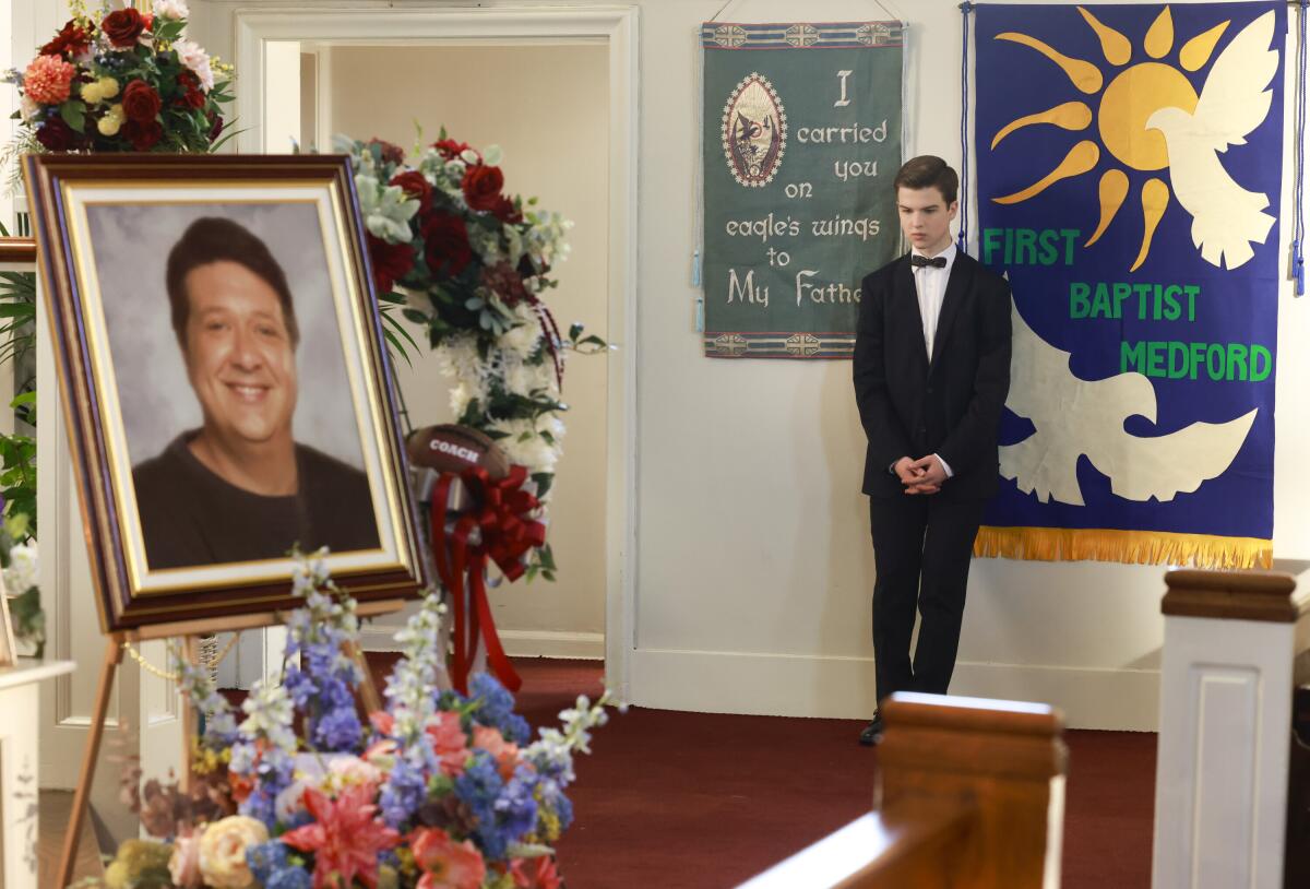  A boy in a black tuxedo looks at a man's photograph on easel surrounded by flowers.