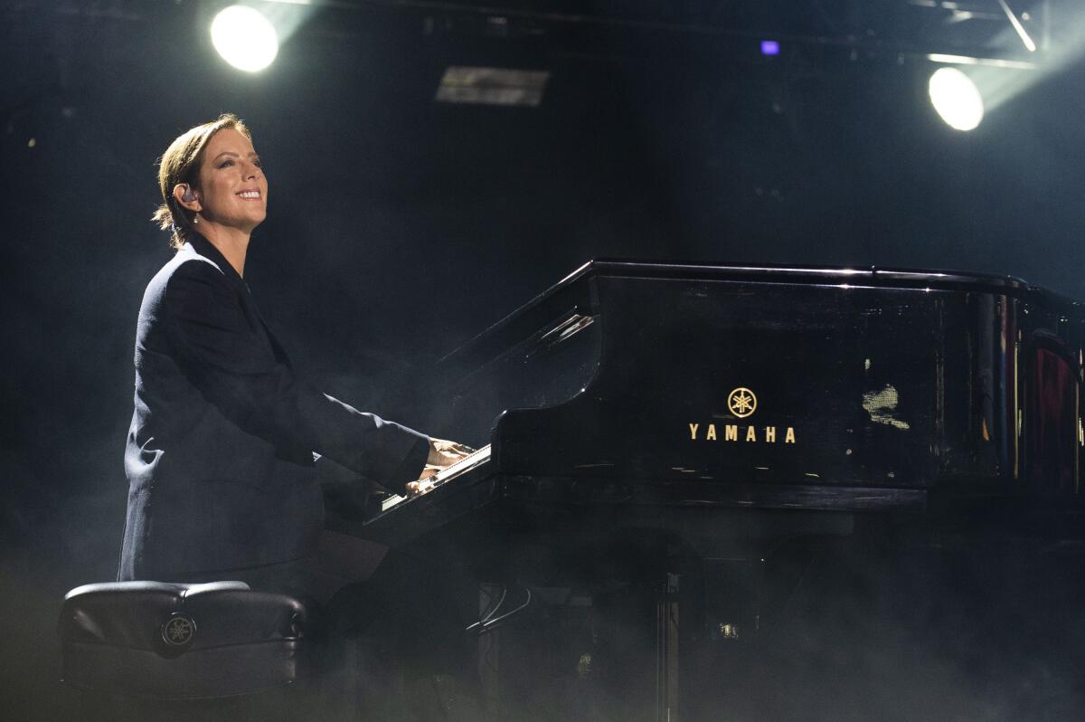 Sarah McLachlan in a black suit sitting at a piano with her hands on the keys. She smiles out to an audience