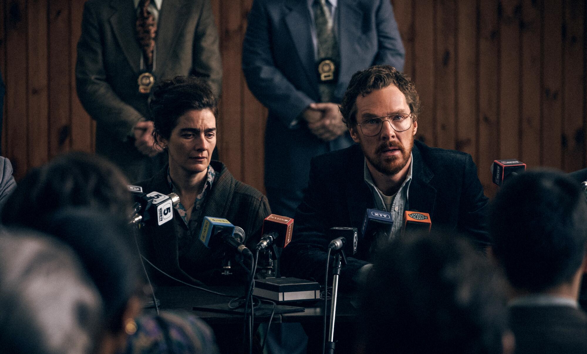 A couple at a table with microphones looking at a crowd of reporters.