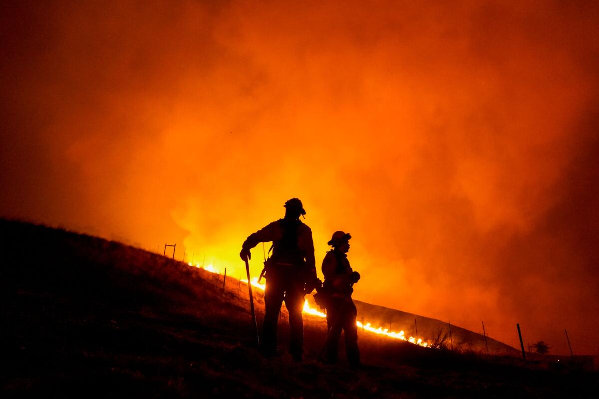 Two firefighters silhouetted against an darkened orange sky as a trail of flames winds up a hillside