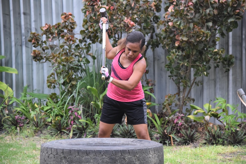 A Papua New Guinean woman wearing a pink singlet and black shorts lifts a huge hammer to hit a tyre.