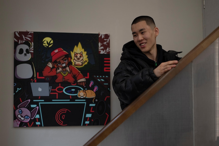 A young Chinese man with short hair stands on a staircase near a painting.