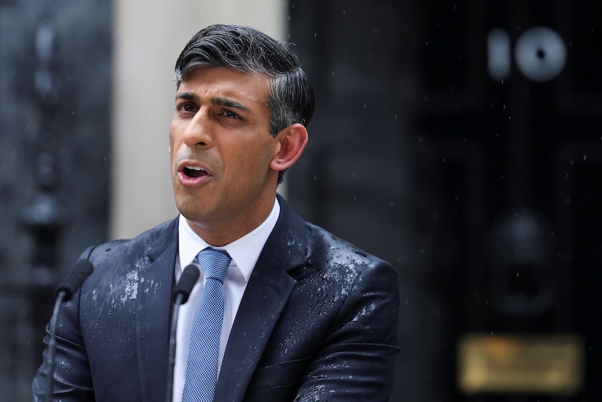 Rishi Sunak stands in a blue suit speaking behind a lectern in the rain in the street