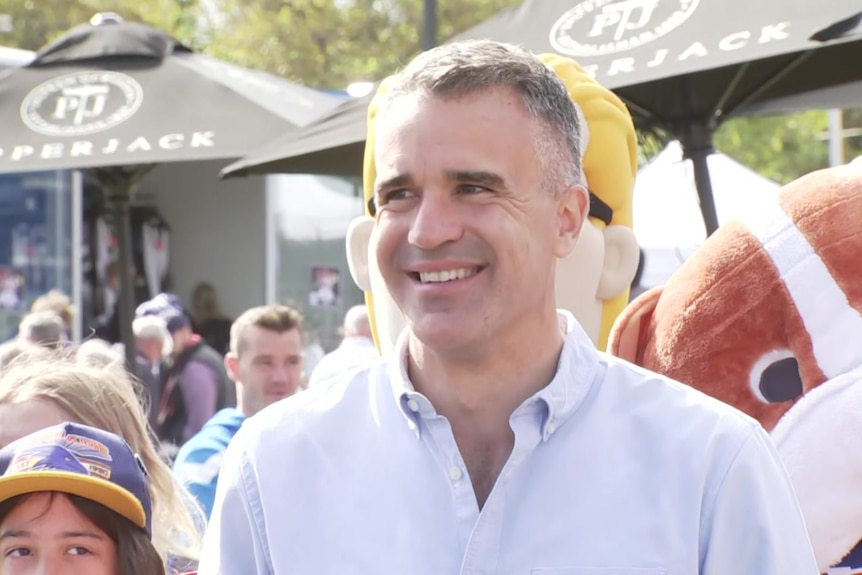 A smiling man in a button up shirt, with a sports mascot and people wandering behind him