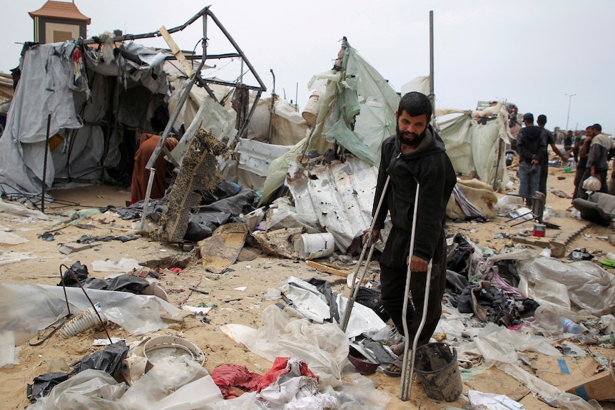 Image of a damaged tent camp. In the middle stands a man with crutches.