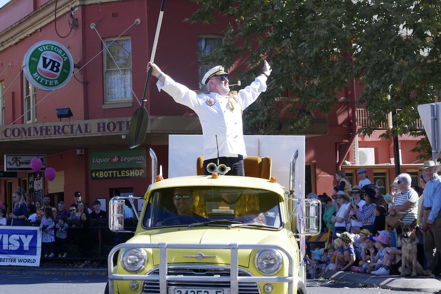 A man stands on a car float with his arms in the air. He wears a white jacket.