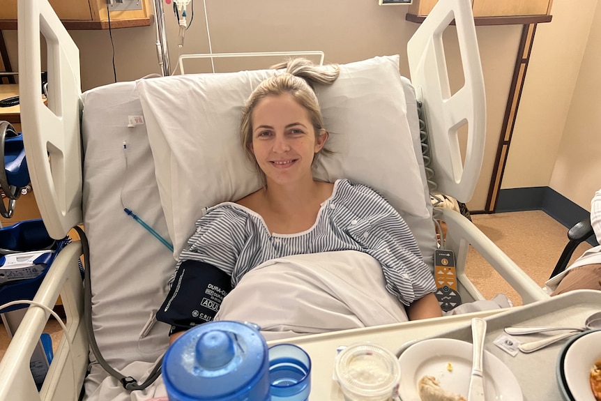 a woman in a hospital gown, sitting up smiling in a hospital bed with a blood pressure cuff on her left arm