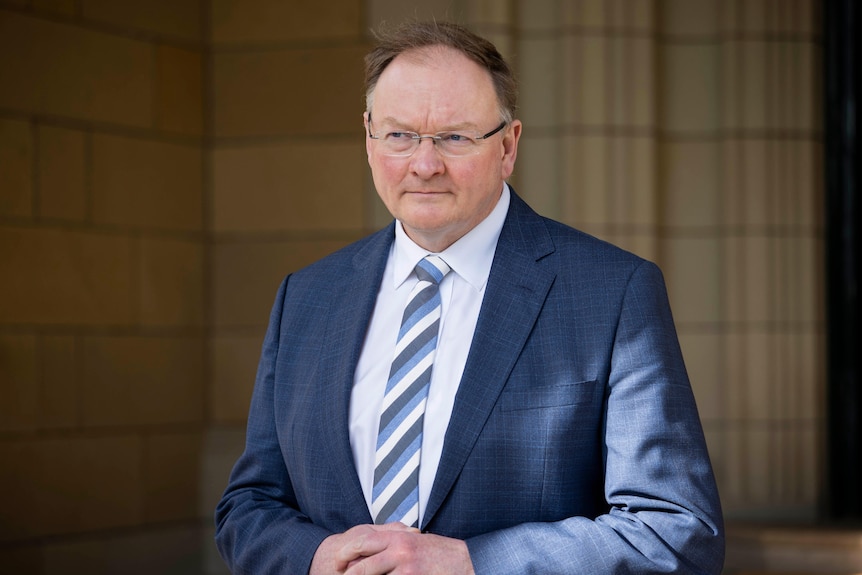 A balding man with glasses wearing a suit stands in a sandstone portico