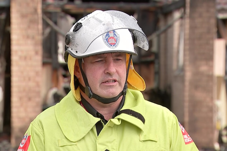 A man dressed in firefighter gear stands in front of a burnt house.