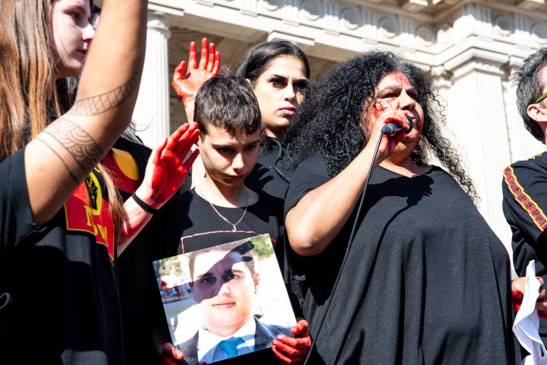 Donnis Kerr. She is at a rally and speaking into a microphone. She has far curly hair and is wearing a black shirt.