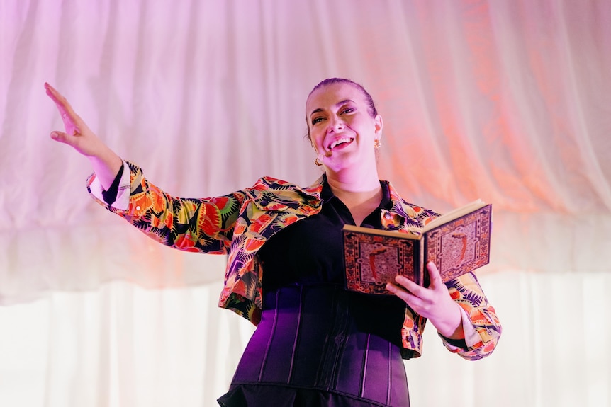 A woman wearing a colourful jacket smiles as she holds a book and gestures with her other hand.