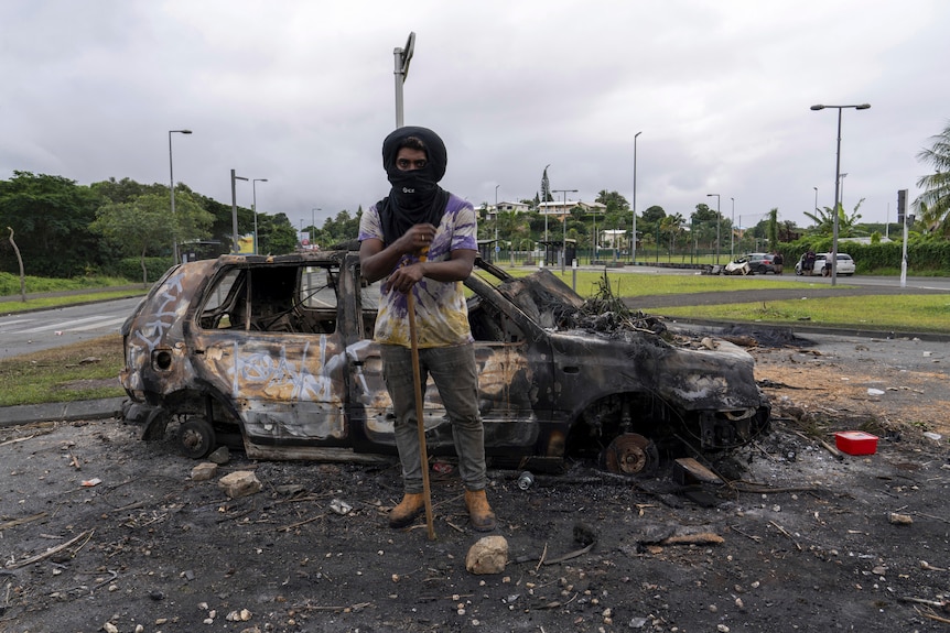 A man in a black hood stands in front of a burnt out car