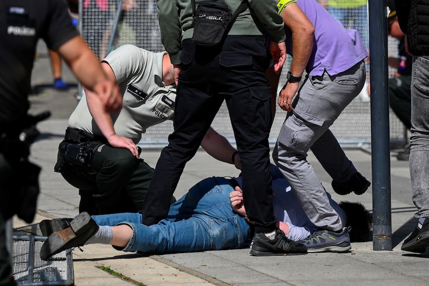 A man wearing jeans is held on the ground by police and bystanders