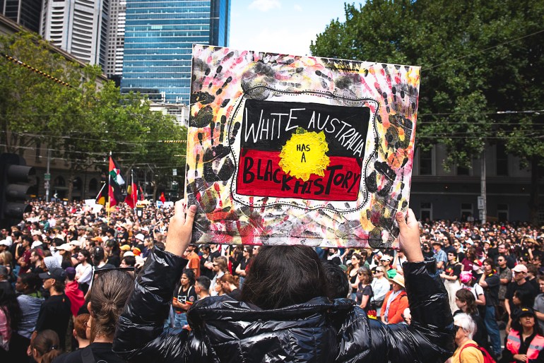 A woman holding up a placard at a rally. It reads White Australia has a Black History' on an Indigenous flag, and there are black handprints across it. There is a sea of peope in front of her.