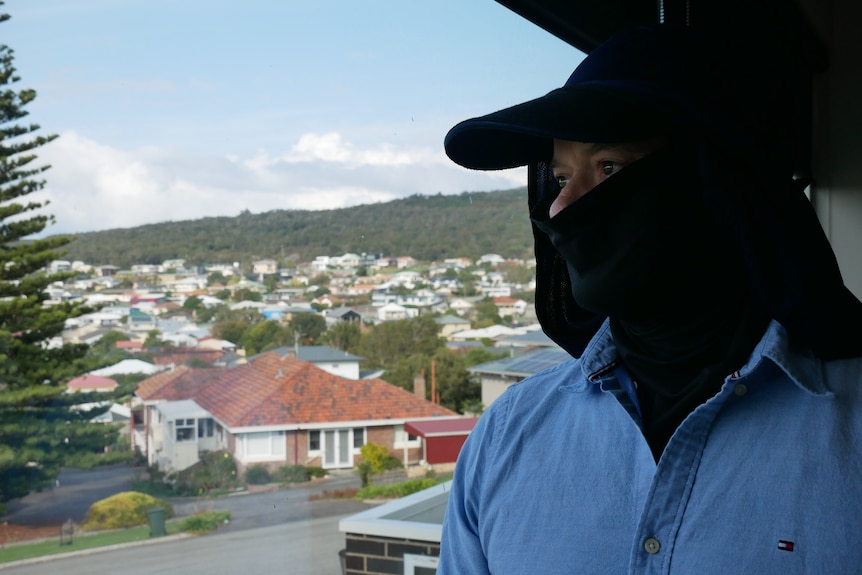 A man stands near a window, wearing a hat and face covering. 