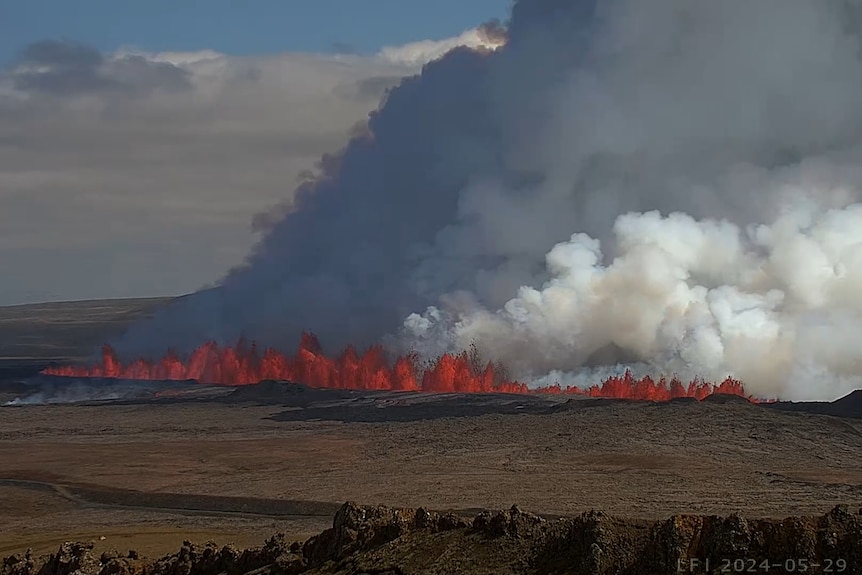 Bright orange lava spews from the ground in a long line, with smoke and ash billowing into the sky