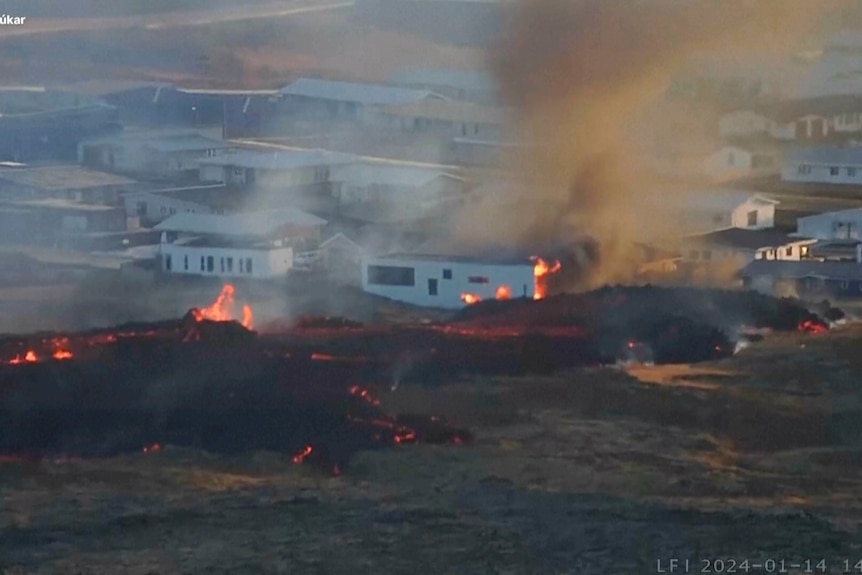 Fire burns on ground in front of a collection of white buildings, with one building on fire.