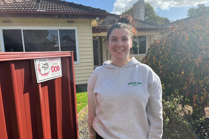 Em is smiling outside her from hate there is a tree and her house in the background, she wears a grey hoodie 