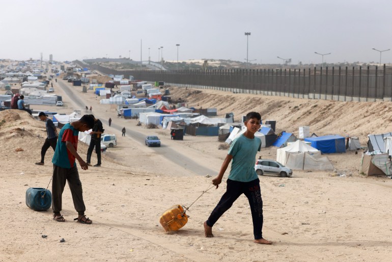 two boys drag water in front of tents in the desert near a border wall