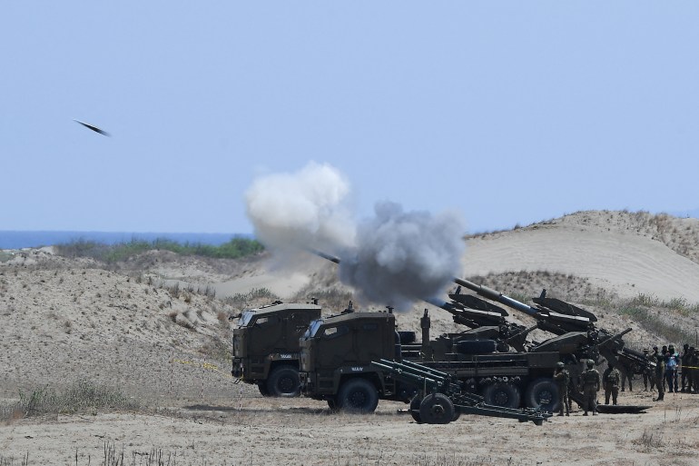 Philippine army personnel fire Autonomous Truck Mounted howitzer system (ATMOS) guns. The weapons are on a beach behind sand dunes. The sea is over the dunes. There are clouds of white smoke. One missile can be seen in the air.