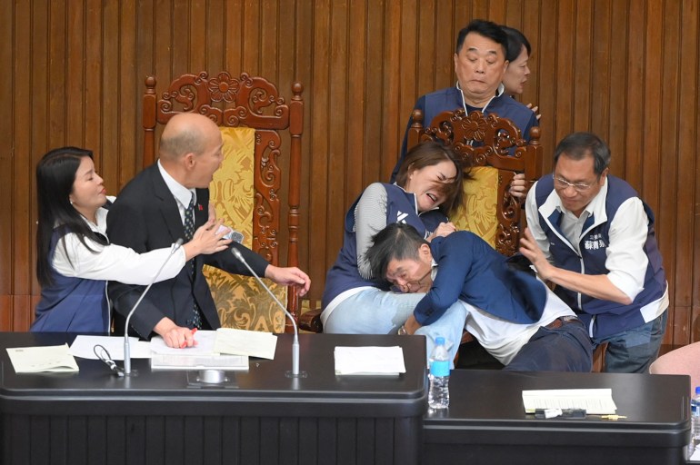 Taiwan legislators brawling in the parliament. A make MP is grabbing a female MP. Others are shouting and pointing. The Speaker is watching.