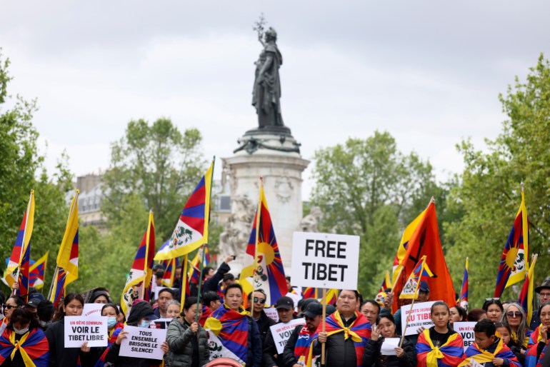 Tibet protesters rally in Paris as Xi Jinping arrives. They have placards reading 'Free Tibet' and are carrying Tibetan flags. There's a statue behind them.