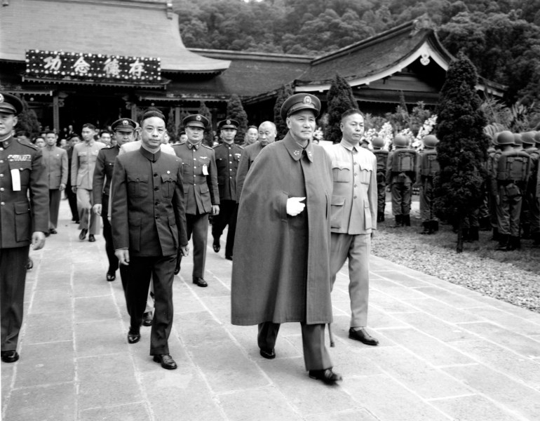 Chiang Kai-shek pictured in 1955. He is wearing a military uniform with a long cape. Others in uniforms are walking behind him. They are leaving a temple.