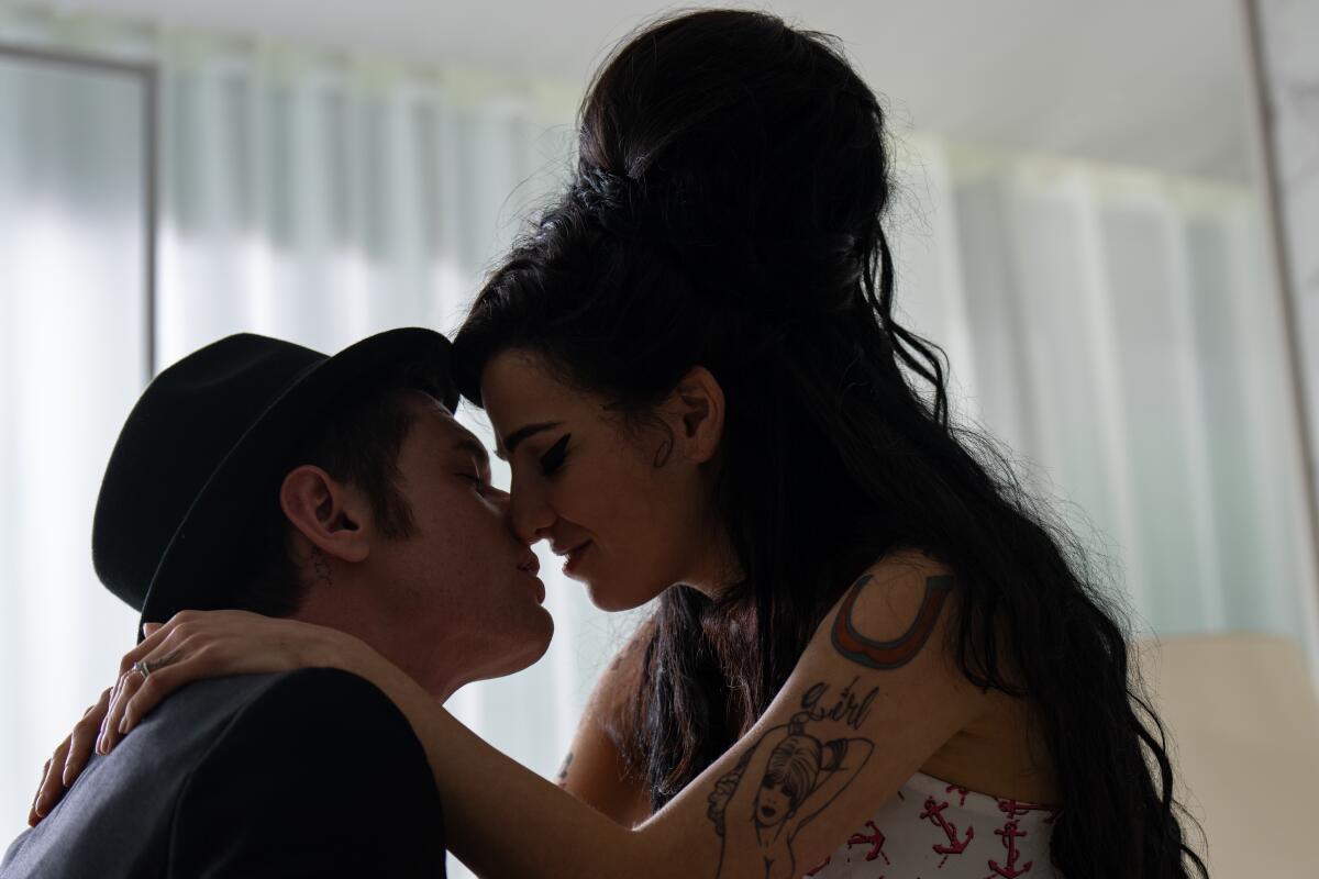 A woman with a big beehive hairdo leans in to kiss a man wearing a hat.