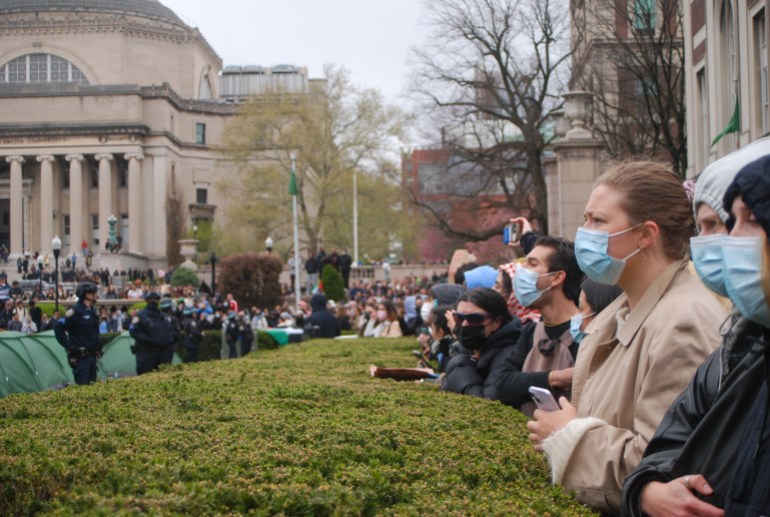 Students in face masks, standing behind a hedge, watch police disband an encampment at Columbia University 