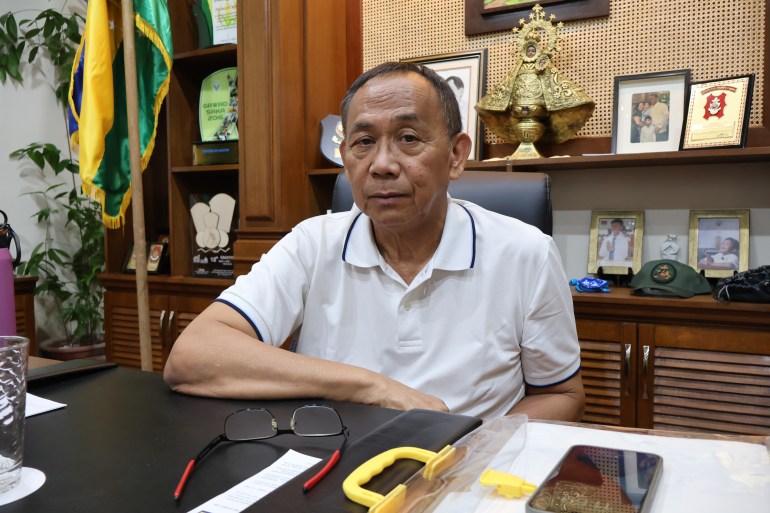 Manuel Mamba sitting at his desk. He is wearing a white polo short and is resting his elbow on the desk and has placed his glasses in front of him. There are family photos and memorabilia on the book case behind him and a yellow and green flag to one side.