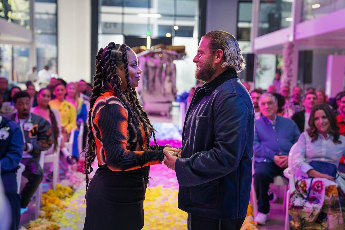 Lauren London and Jonah Hill face each other while holding hands in front of a crowd sitting in pews at a church. 