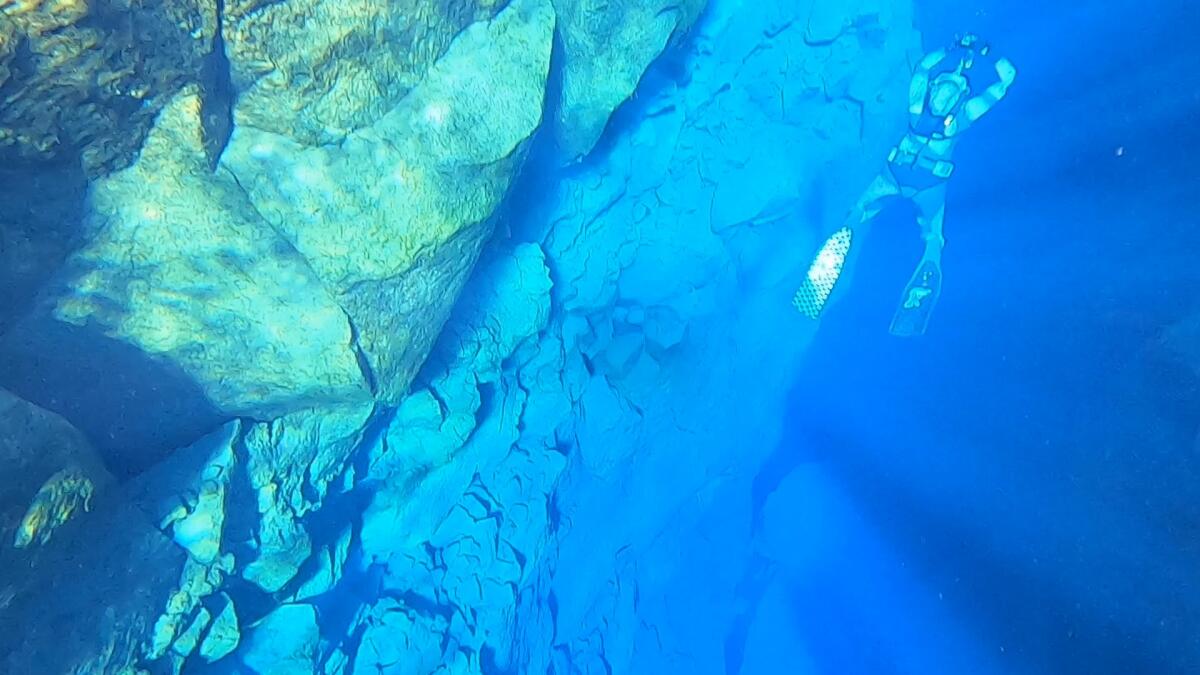 A scuba diver swims past a massive underwater rock formation glowing in blue light.