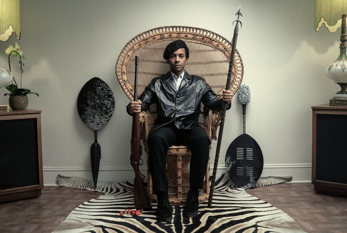 A man in black clothing sitting on a wicker chair holding a rifle and a spear.