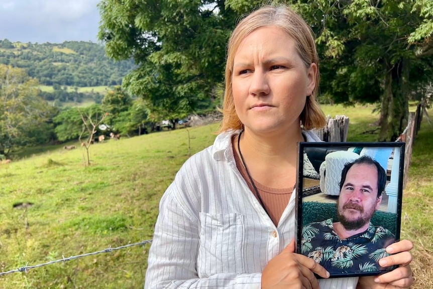 A woman with blonde hair standing in a green paddock, holding a framed photo of a man.