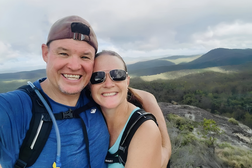 A smiling man and woman in climing gear cuddle up for a selfie on the top of a mountain.