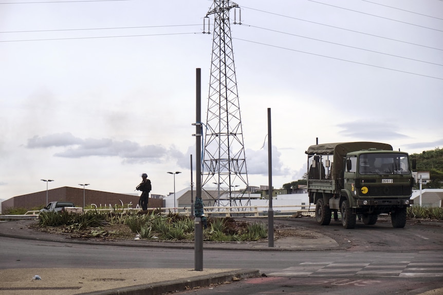 An army van drives slowly along a road with an office in uniform holding a gun walking slowly behind it