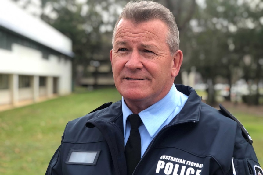 A man in a police jacket stands outside on some grass.