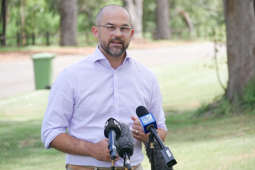 A bespectacled man stands outdoors and speaks to the media.