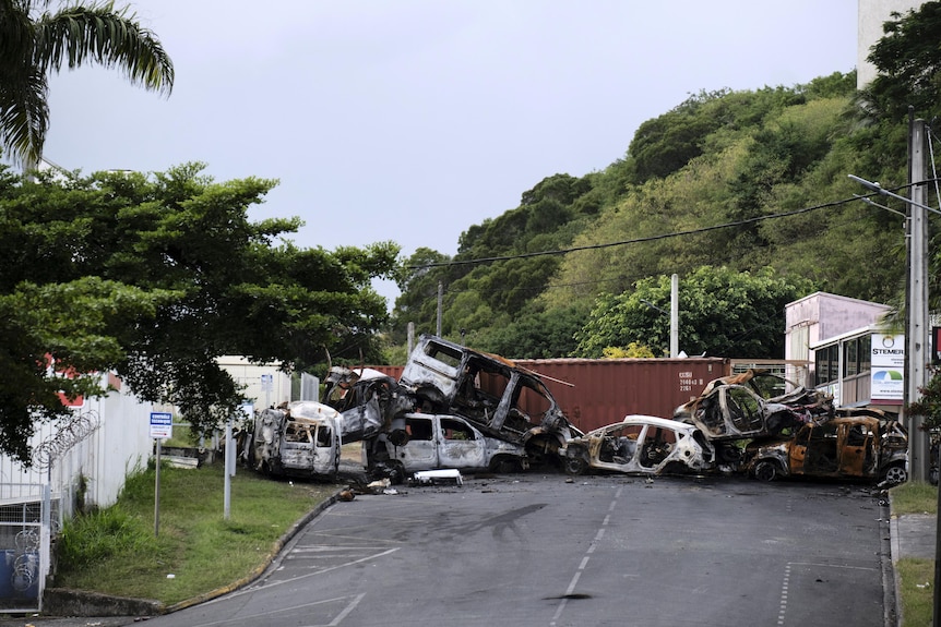 Burnt-out cars sit stacked on top of each other in a street lined by green trees