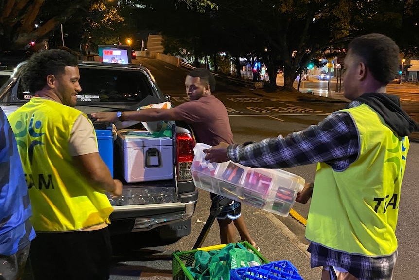 Three men pack boxes and equipment into the back of a ute at night in the city.