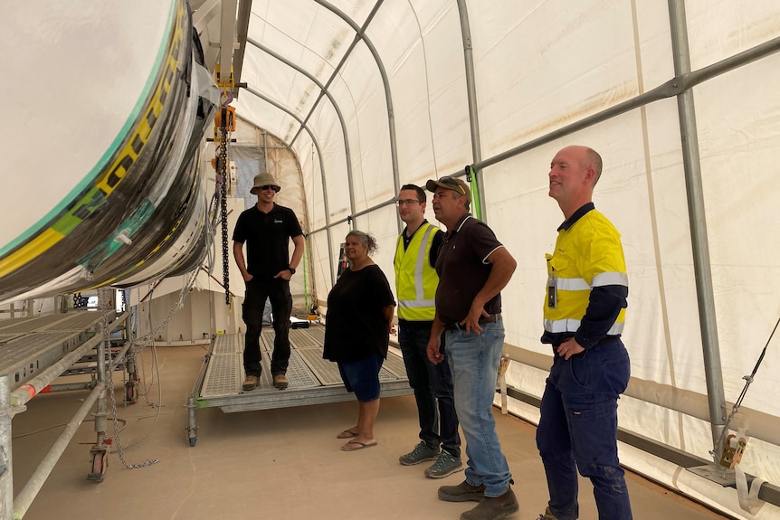 Five people standing on right in large white tent, looking rocket lying on its side, rocket obscured 