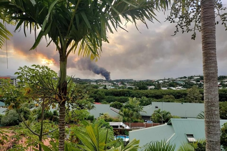 A thick column of black smoke rises from a settlement seen in the distance over water and verdant stretches of land. 