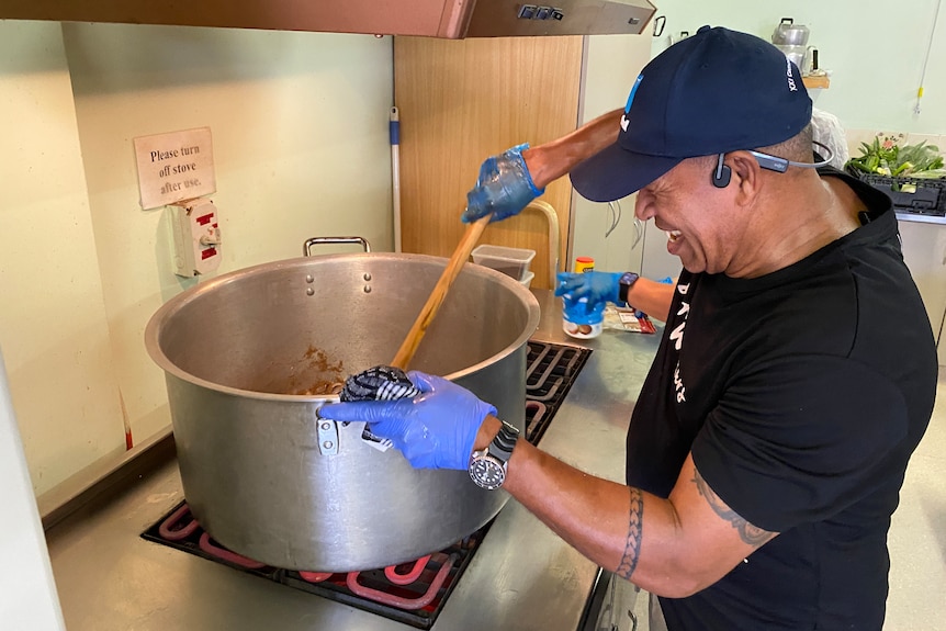 A man in a black shirt and baseball cap smiles while stirring a large pot at a large stove.