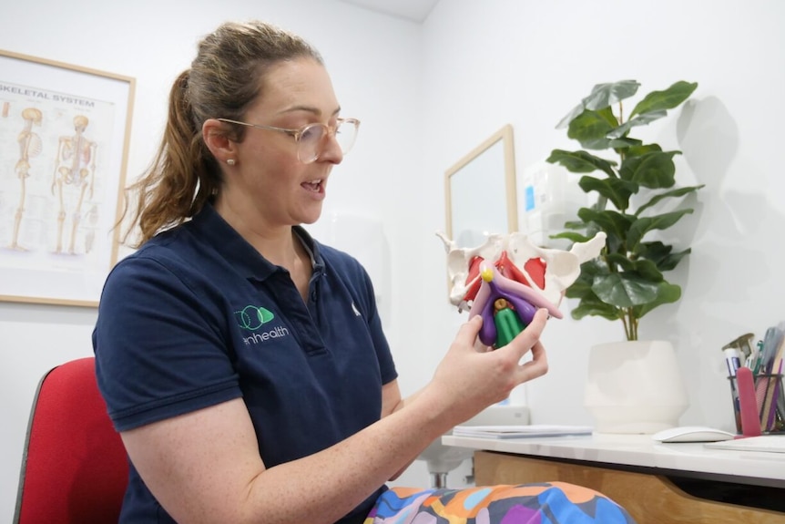 A woman in a doctor's office holding up a model of pelvic and vaginal bones. 