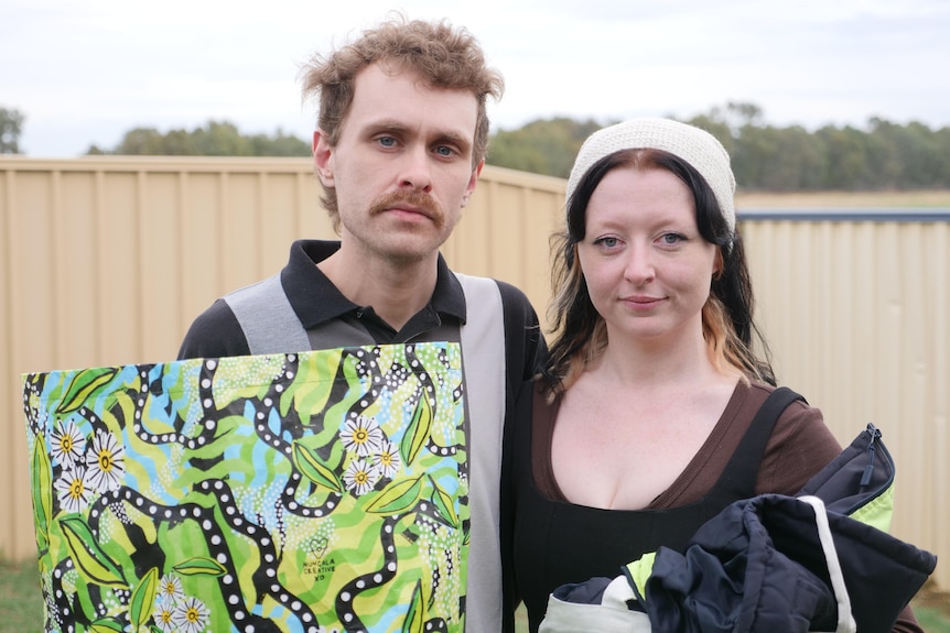 A man and a woman look seriously at the camera, outside under and overcast sky.