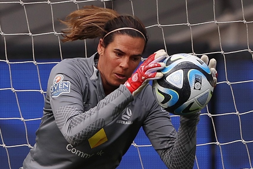 Lydia catches a soccer ball with her gloves hands right near her face.