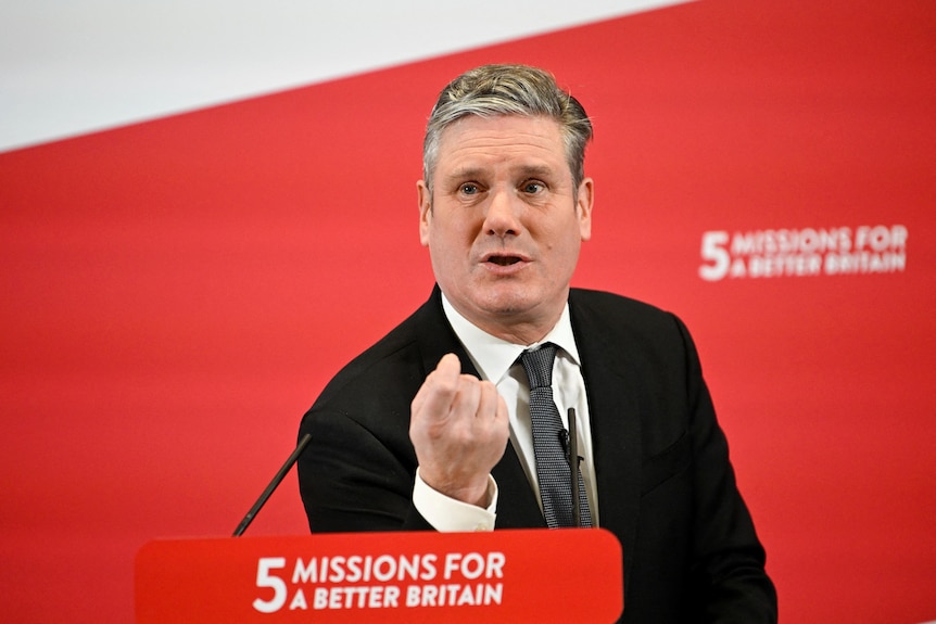 A man stands behind a lectern inside in front of a red banner that says '5 missions for a better future'