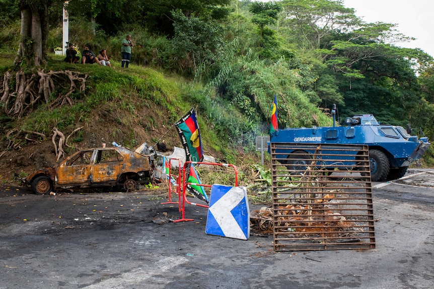 A burnt-out car sits on a road with a Kanak flag hanging from its boot. A gendarme vehicle is parked behind it