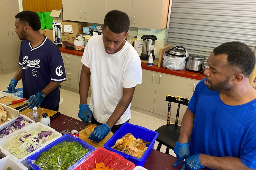 A woman and three men in a kitchen. The men are chopping vegetables and singing.